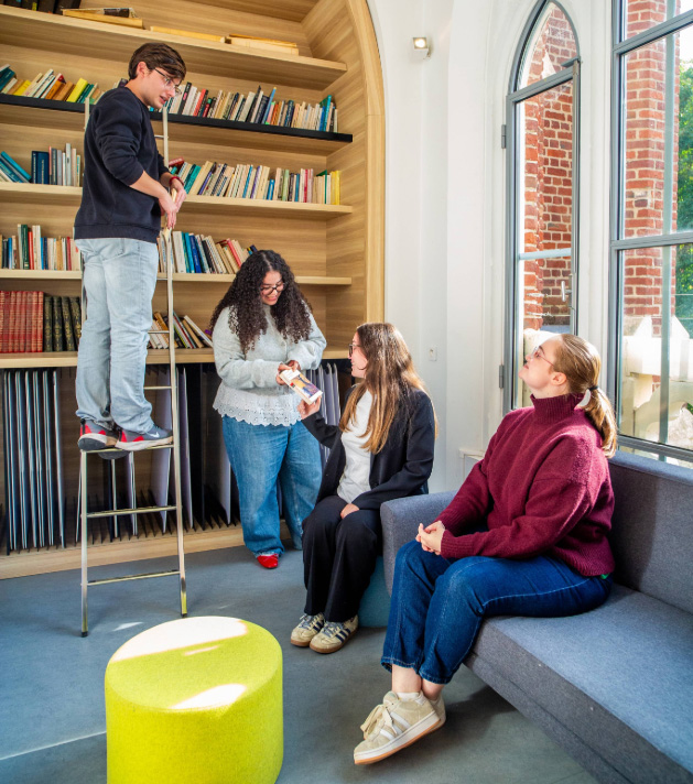 Quatre étudiants discutent dans une bibliothèque moderne et lumineuse. Un jeune homme est debout sur un escabeau devant des étagères de livres en bois courbé. En bas, deux jeunes femmes sont assises sur un canapé gris et une troisième étudiante, debout, leur montre un livre. La pièce est baignée de lumière naturelle provenant d'une grande fenêtre en arche