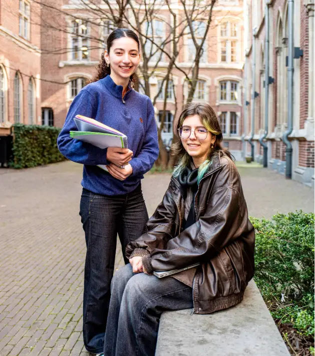 Deux jeunes femmes souriantes posent dans la cour intérieure d'un bâtiment ancien en briques rouges. L'une est debout, vêtue d'un pull bleu et d'un pantalon noir, tenant plusieurs cahiers colorés contre elle. L'autre est assise sur un muret en pierre, portant un blouson en cuir marron, des lunettes et un pantalon gris