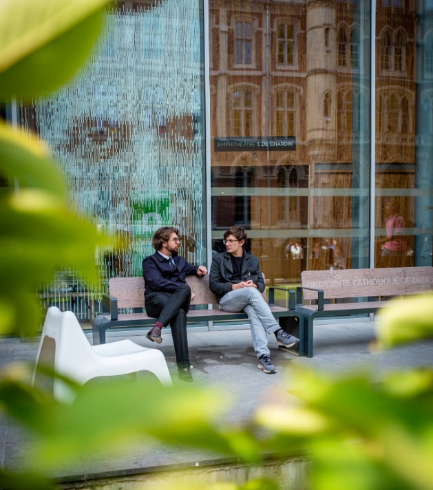 Deux jeunes hommes discutent, assis sur un banc devant l'entrée moderne d'un bâtiment universitaire. En arrière-plan, on aperçoit une grande baie vitrée reflétant une architecture ancienne en briques, avec une inscription indiquant « Amphithéâtre T. de Chardin » et « Université Catholique de Lille ». La photo est prise à travers un feuillage vert flou au premier plan, créant un effet de cadre naturel