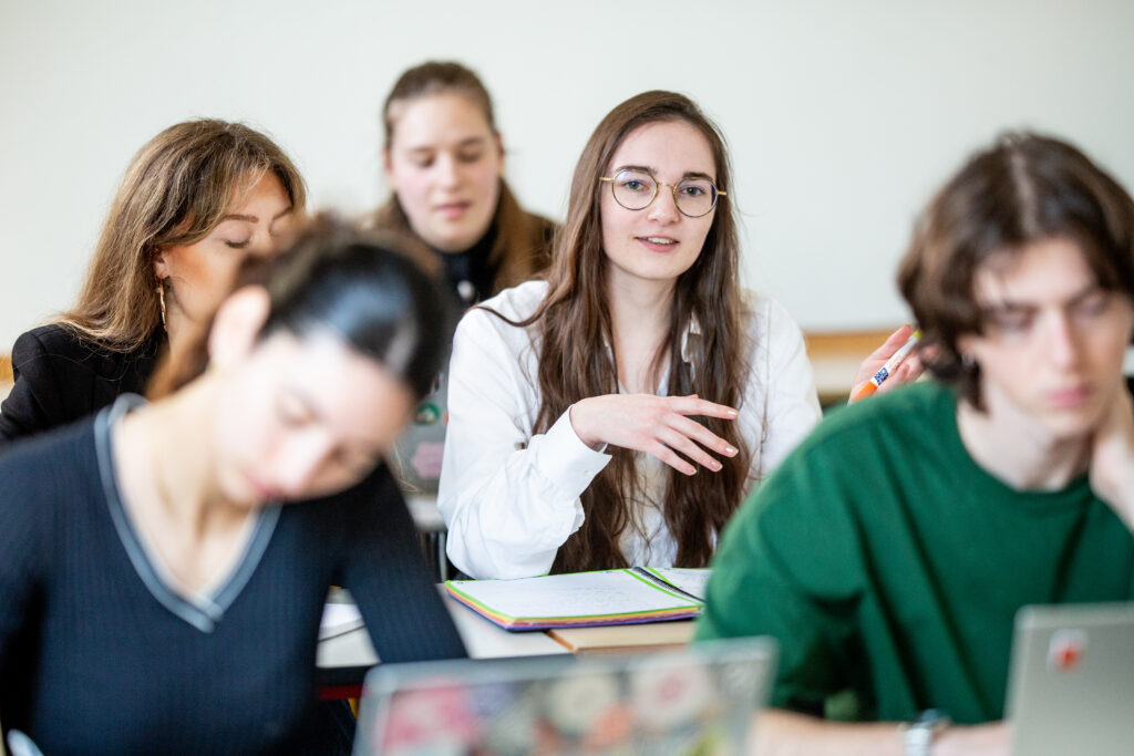 Un groupe d'étudiants en salle de cours, avec une jeune femme au centre qui participe activement à la discussion.