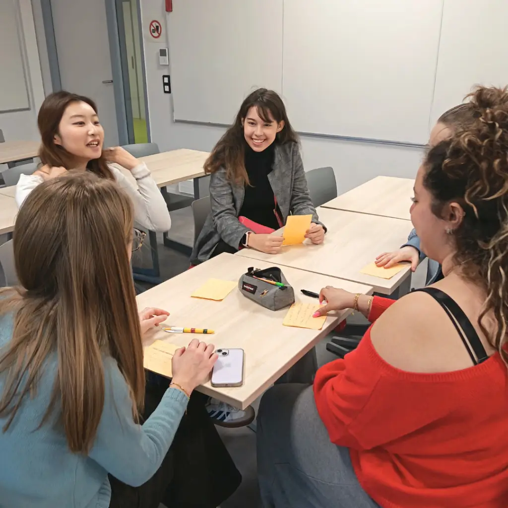 Un groupe de cinq étudiantes et étudiants réunis autour d'une table de cours pour un travail de groupe. Ils discutent avec le sourire et utilisent des notes autocollantes (post-its) jaunes pour organiser leurs idées. L'atmosphère est collaborative et dynamique.