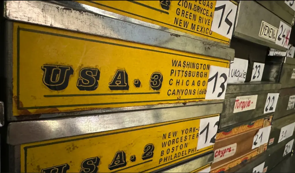 Gros plan sur des boîtes d'archives métalliques empilées. Les boîtes sont jaunes avec des inscriptions noires. On lit distinctement "USA 3" avec les villes de Washington, Pittsburgh, Chicago et Canyons, ainsi que "USA 2" avec New York, Worcester, Boston et Philadelphie. Des étiquettes numérotées (12, 13, 14) sont collées sur le côté droit