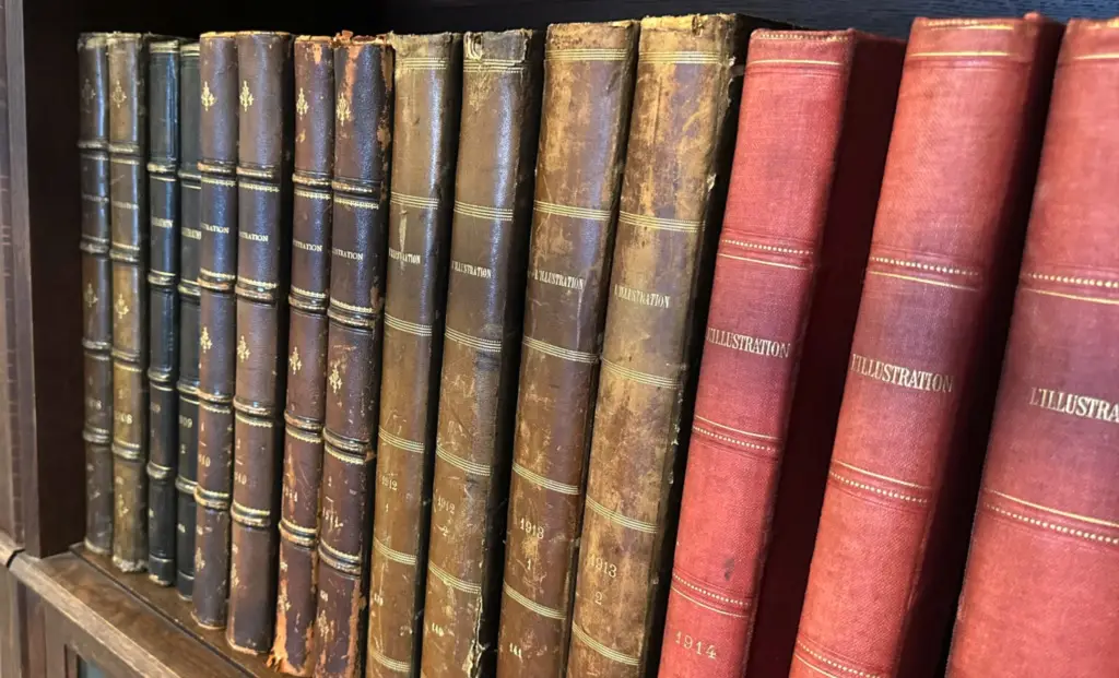 Une rangée de vieux livres reliés alignés sur une étagère en bois. Les reliures sont en cuir usé, passant du noir au brun foncé, puis au brun clair, pour finir par quatre volumes d'un rouge vif sur la droite. Le mot "ILLUSTRATION" est inscrit en lettres dorées sur le dos de plusieurs ouvrages, avec certaines dates visibles comme "1913" et "1914