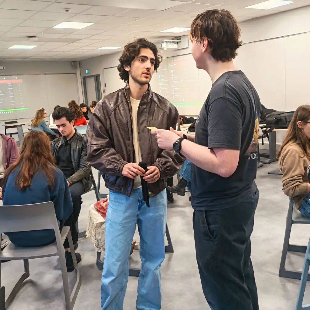 Deux jeunes hommes discutent debout dans une salle de classe universitaire. L'un, portant un blouson en cuir marron et un jean bleu, écoute attentivement l'autre, vêtu d'un t-shirt noir, qui semble lui présenter un petit document ou une carte. D'autres étudiants sont assis en arrière-plan
