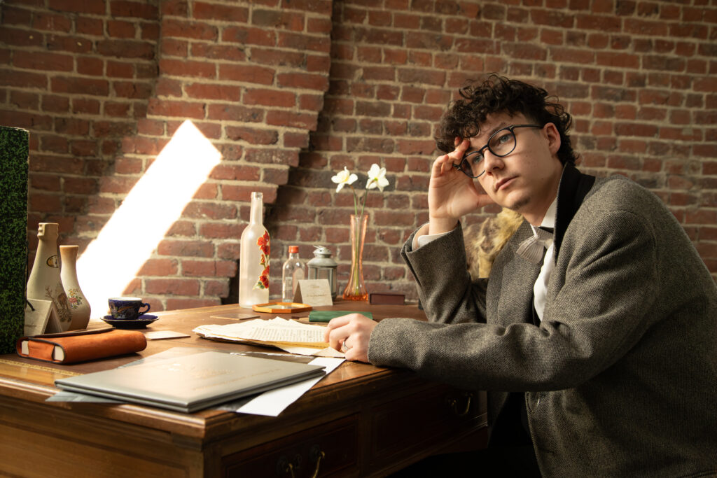 Portrait d'un homme à lunettes en veste grise, assis à un bureau d'étude vintage devant un mur de briques