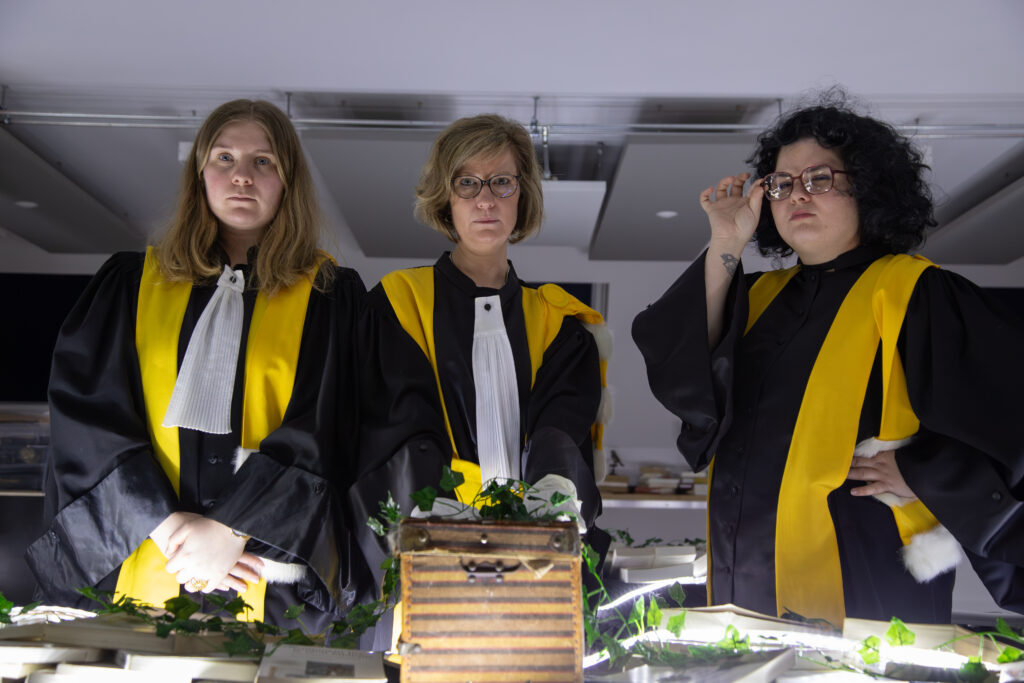 Trois femmes en tenues académiques solennelles posant derrière un coffret ancien.