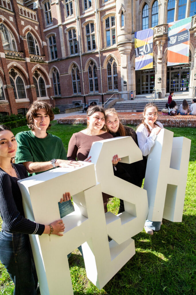 Quatre jeunes étudiants et étudiantes posent avec sourire derrière de grandes lettres blanches en volume formant le mot « FLSH ». Ils se trouvent sur une pelouse, devant un bâtiment universitaire historique en briques rouges avec des fenêtres ogivales. À l'arrière-plan, on aperçoit des drapeaux colorés et le logo de l'Université Catholique de Lille.